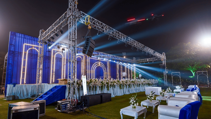 An outdoor event space at Garh Govind featuring a metal truss stage structure with blue backdrops and tables illuminated by spotlights.