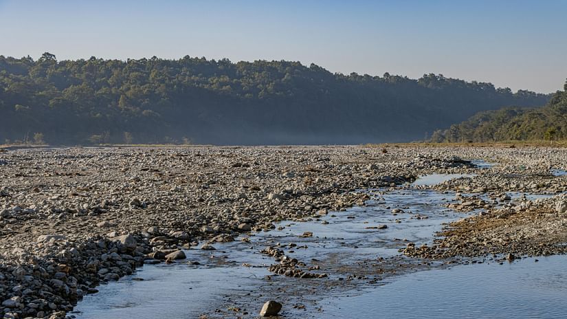 A wide shot of a rocky riverbed with a small stream flowing through it, backed by distant hills under a clear sky.