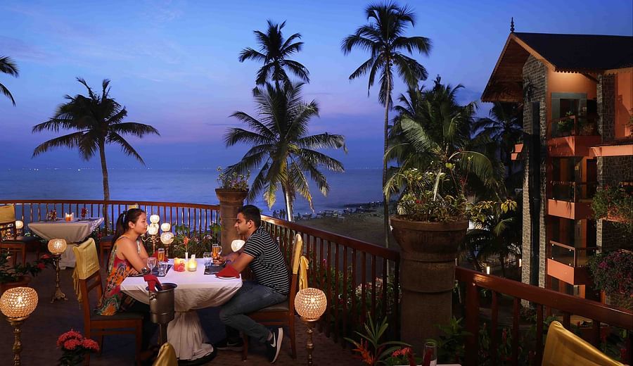 A couple enjoys a romantic candlelit dinner on a balcony overlooking the ocean at dusk, surrounded by palm trees at Uday Samudra Leisure Beach Hotel.