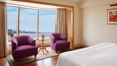 Seating area of a spacious bedroom featuring 2 chairs with a small table beside a curved window overlooking the sea outside.