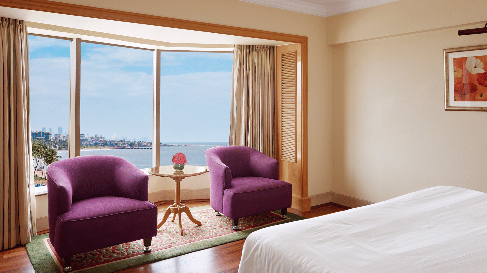 Seating area of a spacious bedroom featuring 2 chairs with a small table beside a curved window overlooking the sea outside.