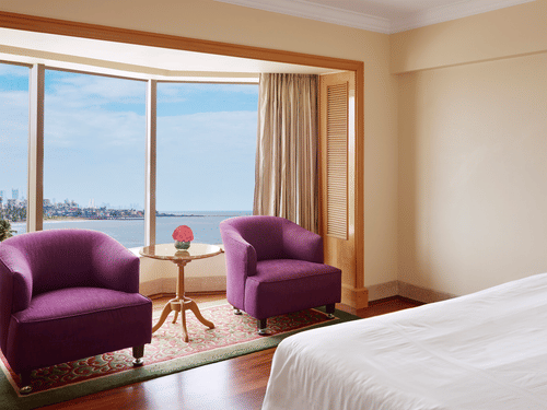 Seating area of a spacious bedroom featuring 2 chairs with a small table beside a curved window overlooking the sea outside.