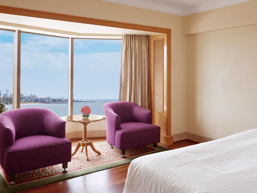 Seating area of a spacious bedroom featuring 2 chairs with a small table beside a curved window overlooking the sea outside.