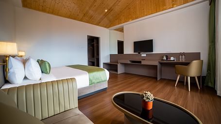 Spacious hotel room with a large bed featuring green accents, a wooden ceiling, cosy seating area with a coffee table, work desk with a TV, and wooden flooring - Yashshree Pelling Chekhim Retreat.