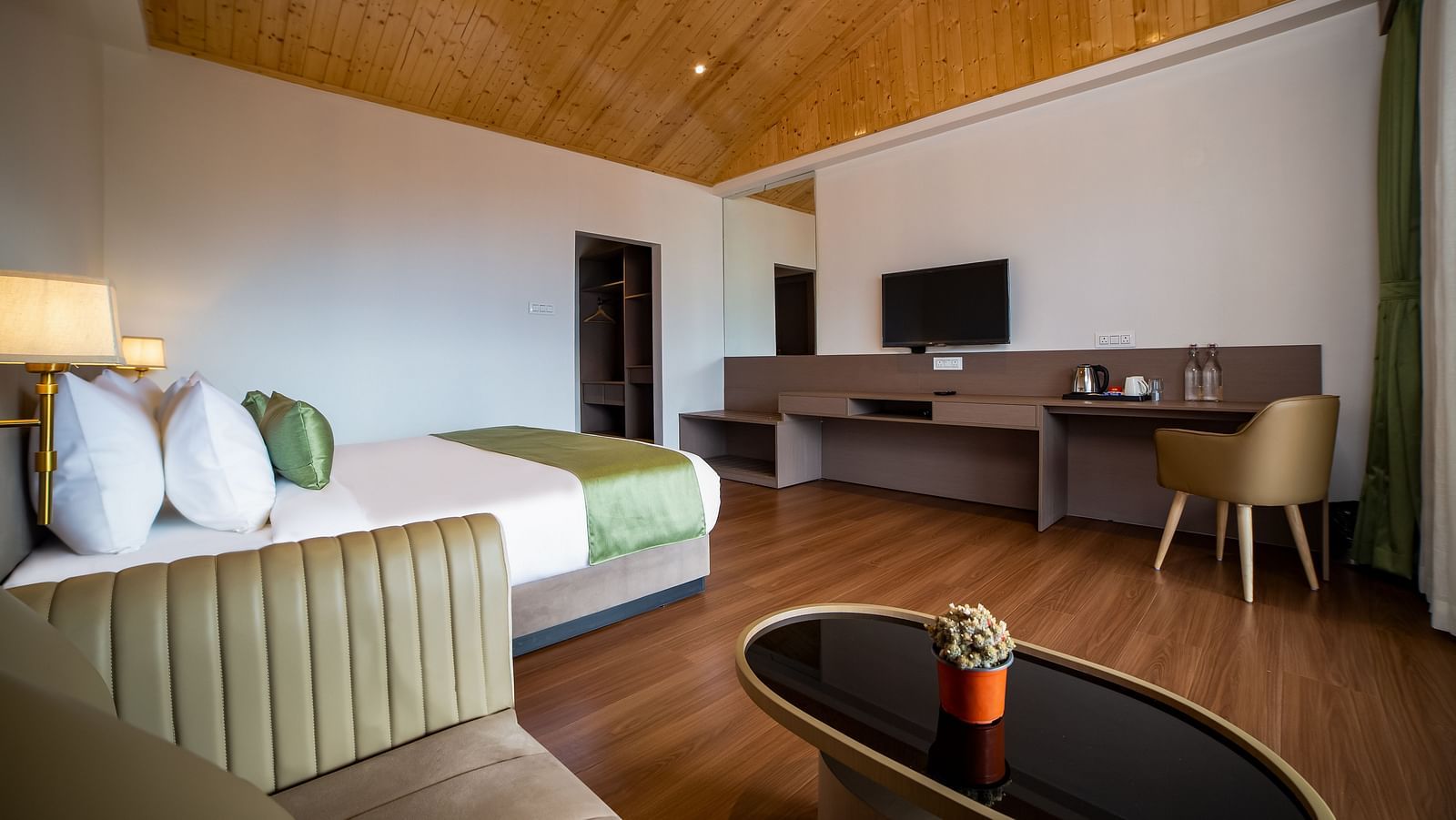 Spacious hotel room with a large bed featuring green accents, a wooden ceiling, cosy seating area with a coffee table, work desk with a TV, and wooden flooring - Yashshree Pelling Chekhim Retreat.