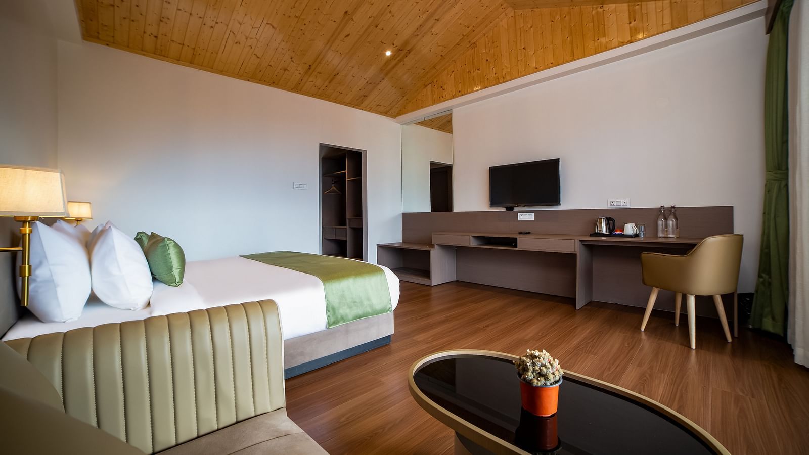 Spacious hotel room with a large bed featuring green accents, a wooden ceiling, cosy seating area with a coffee table, work desk with a TV, and wooden flooring - Yashshree Pelling Chekhim Retreat.