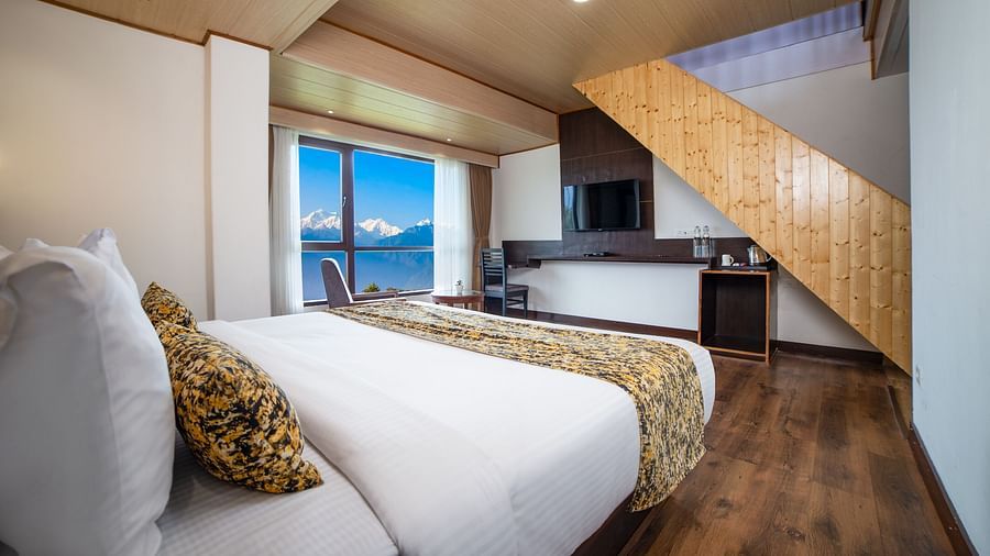 Sumitel hotel's bedroom with a double bed, white linens, yellow accent pillows, a wooden floor, and a partial view of a kitchen space