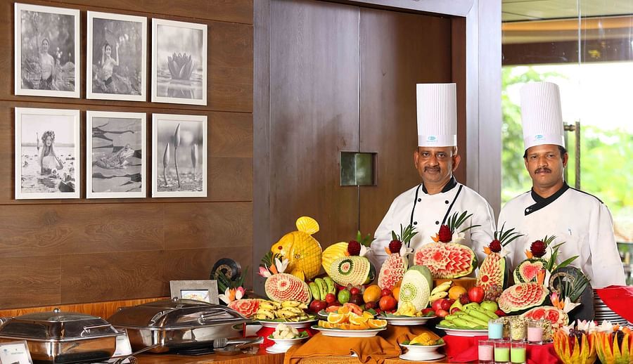 Two chefs in white uniforms and tall hats stand behind a buffet table displaying elaborately carved fruits and vegetables, including watermelons, pineapples, and other coluorful produce - UDS Group of Hotels.