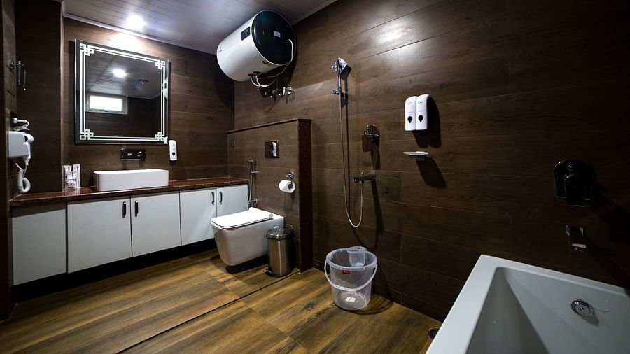 Modern hotel bathroom with wooden walls and flooring, featuring a wall-mounted toilet, white vanity with a sink and mirror, overhead water heater, shower area with dispensers, and a transparent bucket on the floor - Yashshree Lachen.