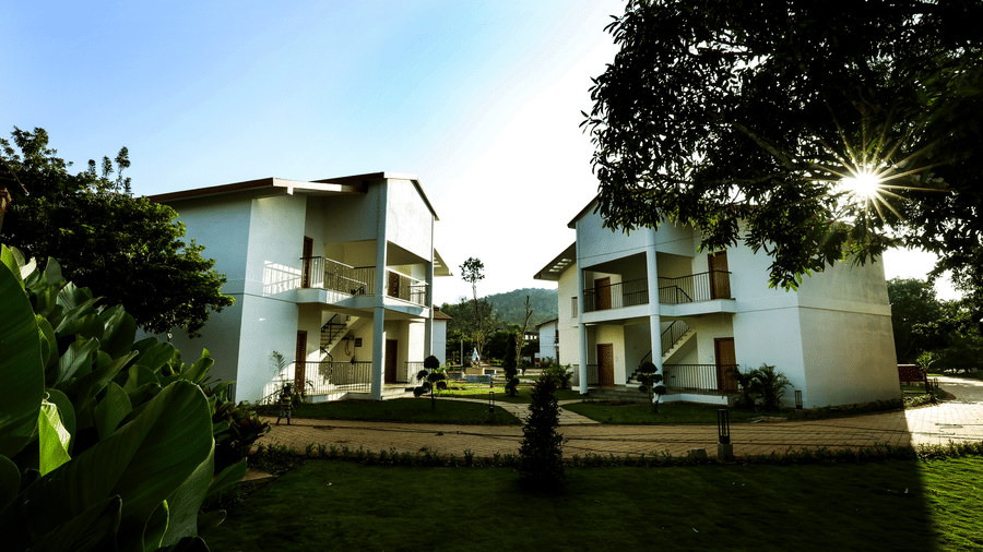 An exterior view of white multi-storey resort buildings surrounded by green lawns and trees under a clear sky | Lords Sahyadri Resort, Shivamogga