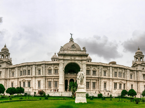 A wide long shot of the Victoria memorial showcasing its British architecture and green gardens during a cloudy day.