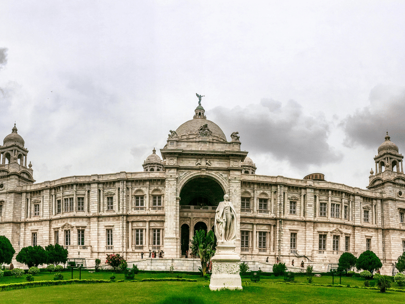 A wide long shot of the Victoria memorial showcasing its British architecture and green gardens during a cloudy day.