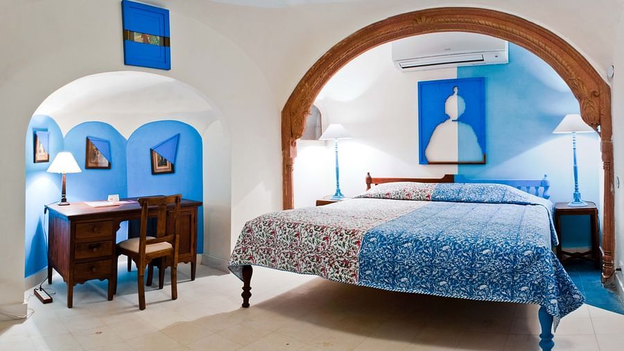 A hotel room with a bed featuring a patterned bedspread, bedside tables with lamps, a desk with a chair in an alcove, and arched architectural details with artwork at Tijara Fort-Palace - 19th Century, Alwar.