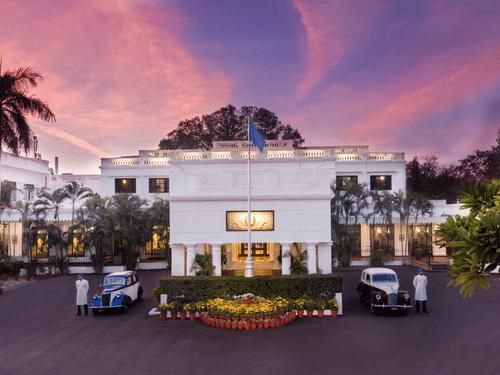 Front view of Jehan Numa Palace in Bhopal at dusk, showcasing the heritage white façade, palm-lined entrance, and vintage cars.