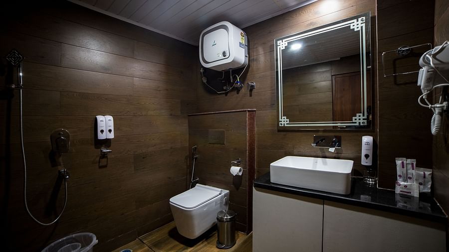 Modern hotel bathroom with dark wooden-style walls, a wall-mounted toilet, a walk-in shower, a countertop sink with a large mirror, and amenities including a hairdryer and water heater - Yashshree Lachen.