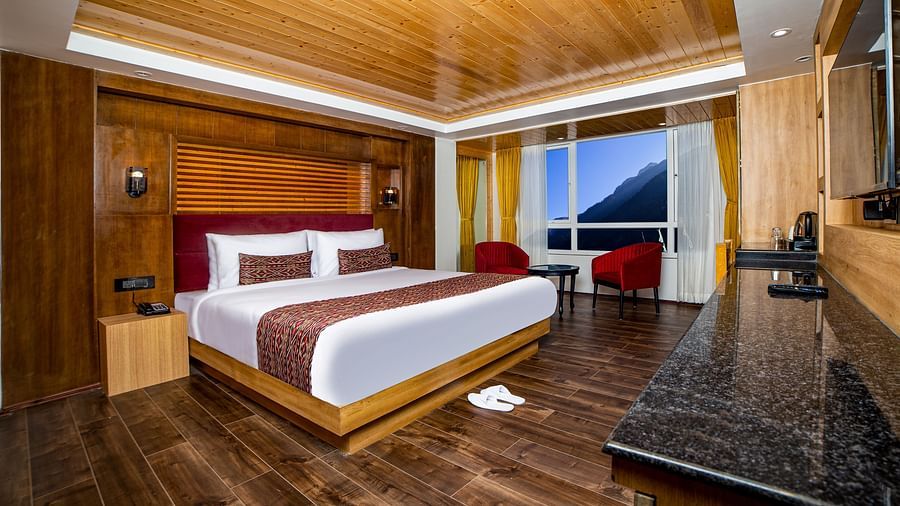 Spacious hotel room with a large bed featuring white linens and red accents, wooden flooring and ceiling, two red chairs near a large window with mountain views, and a black granite countertop along one wall - Yashshree Lachen.