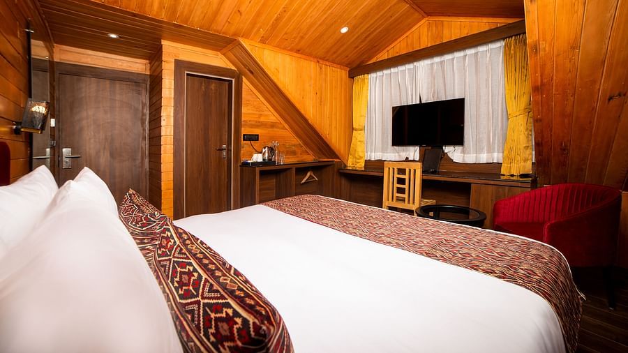 Cosy hotel room with angled wooden ceiling and walls, featuring a large bed with white linens and patterned red pillows, a flat-screen TV mounted above a wooden desk with a chair, a red armchair by the window, and a wooden door in the background - Yashshre