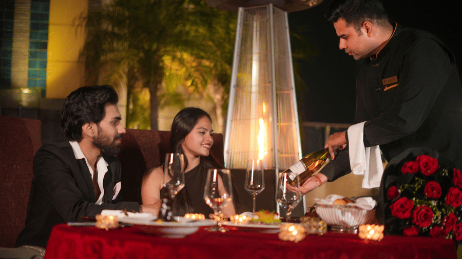 A server standing beside a table placing food for a seated couple at an outdoor dining area at The Suryaa, New Delhi.