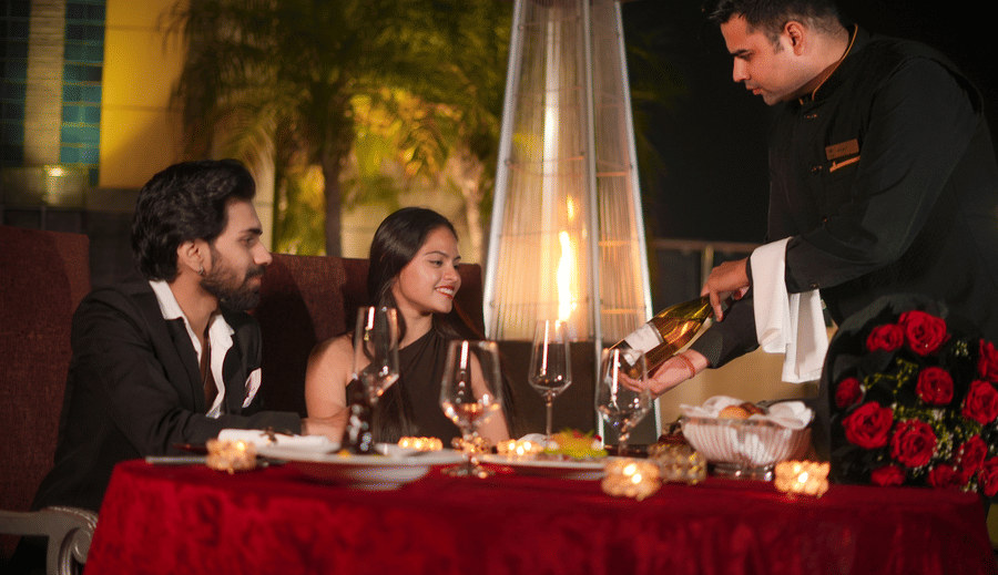 A server standing beside a table placing food for a seated couple at an outdoor dining area at The Suryaa, New Delhi.