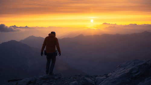 A mountain viewpoint with a yellow coloured sky in the background and a woman standing on the cliff.