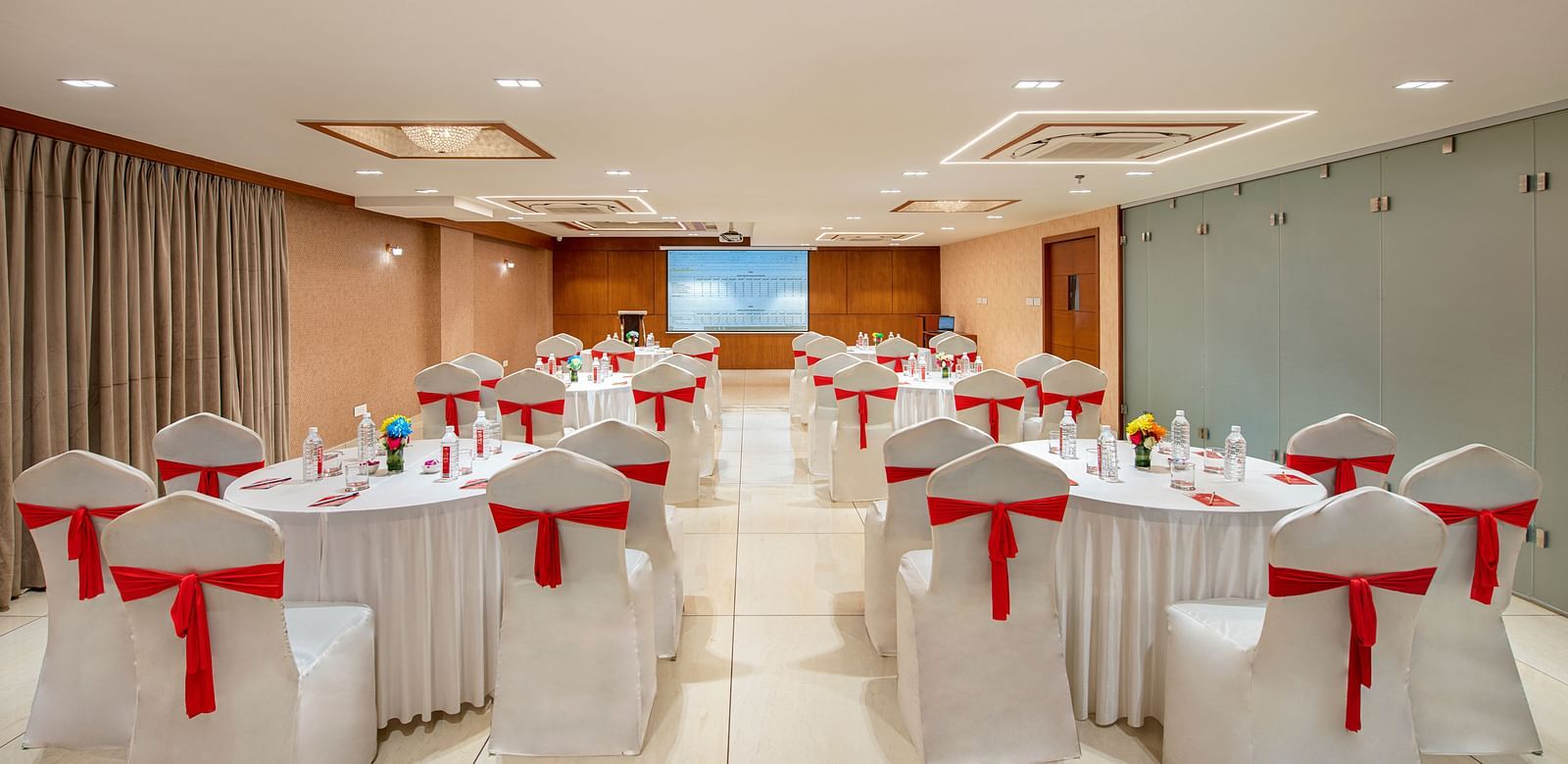 A banquet hall with multiple round tables, each set with  linens and red chair sashes - SK Lords Inn Ahmedabad