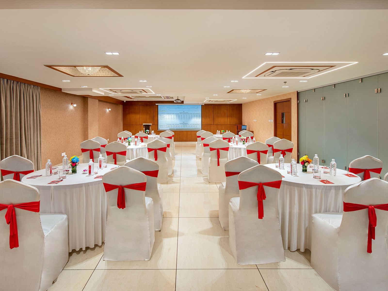 A banquet hall with multiple round tables, each set with  linens and red chair sashes - SK Lords Inn Ahmedabad