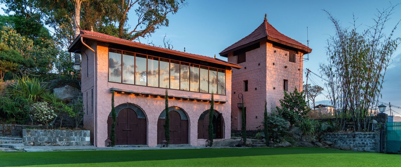  A unique, pink-hued building with a tower, large windows, and arched doorways, situated on a grassy lawn with trees and a bamboo grove.
