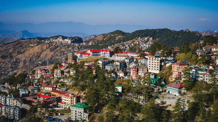 A panoramic view of the hillside city of Shimla, Himachal Pradesh, with numerous buildings nestled among green trees and mountains