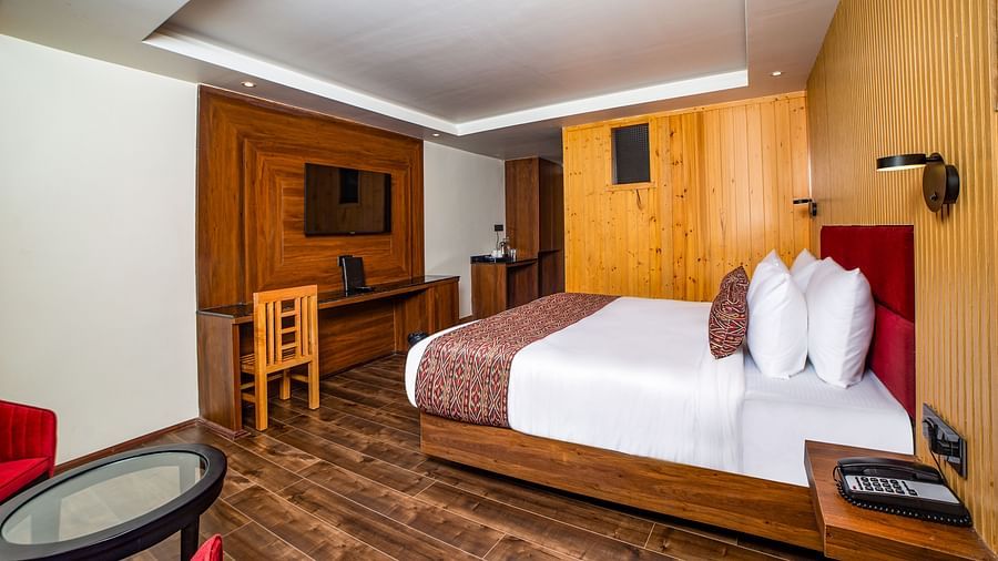 Stylish hotel room featuring a neatly made bed with a red headboard and decorative pillows, wooden flooring and paneling, a work desk with a mirror, and a cosy sitting area with a glass-top table and red chairs - Yashshree Lachen.