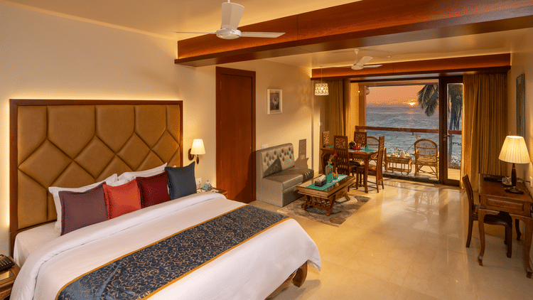  Lounge Seaview Suite at Uday Samudra Leisure Beach Hotel featuring a plush king size bed with colourful pillows, a seating area, and a balcony overlooking the sea at sunset