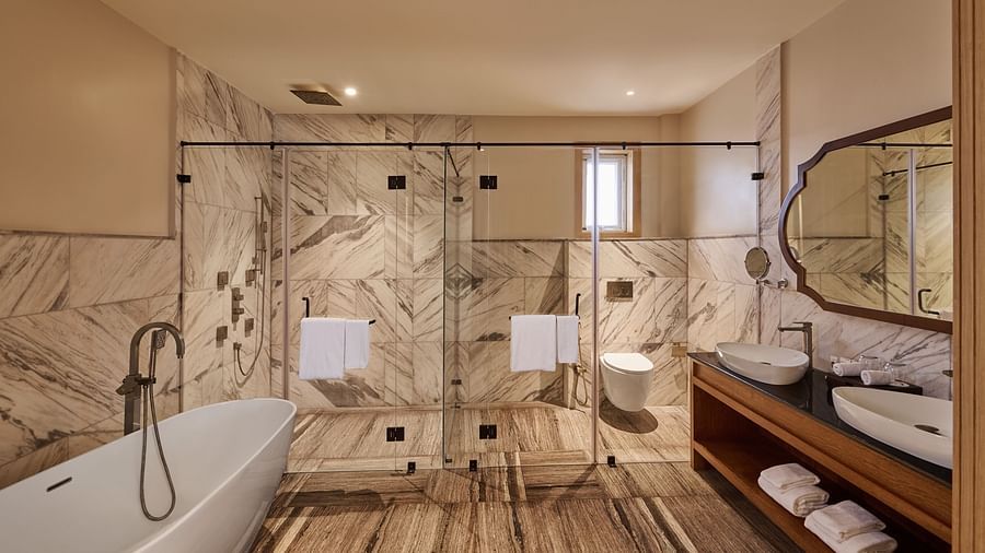 Luxurious bathroom with marble walls and flooring, a freestanding bathtub, glass-enclosed shower and toilet area, double vessel sinks on a wooden vanity with open shelving, and a large decorative mirror - Voyage The Solitaire Bliss.