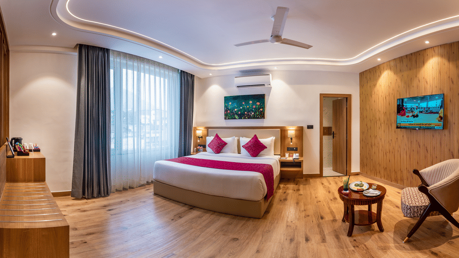 A well-lit bedroom at Lords Inn Katra featuring a king size bed, a wall-mounted television screen, and a wooden chair next to a coffee table.