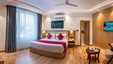 A well-lit bedroom at Lords Inn Katra featuring a king size bed, a wall-mounted television screen, and a wooden chair next to a coffee table.