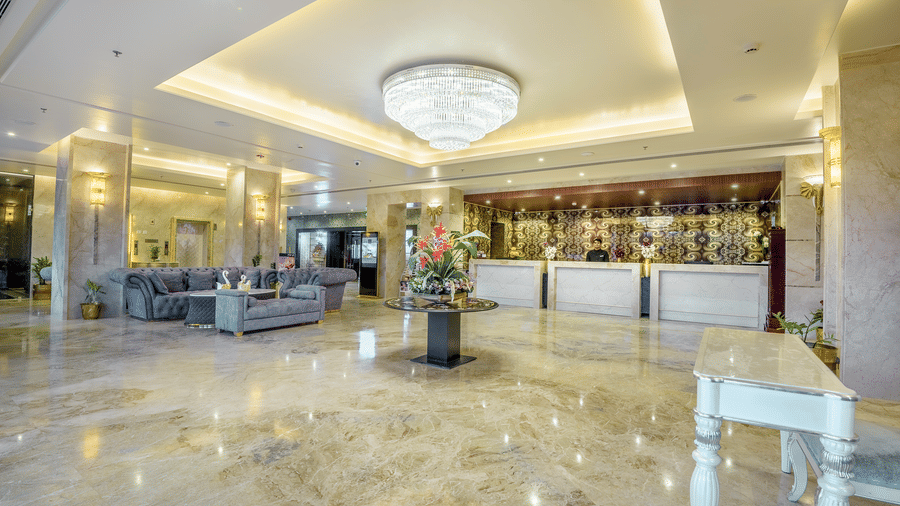 A bright, wide-angle shot of a grand hotel reception area with polished stone flooring, elegant floral displays, and a long service desk at Golden Tulip Kukas, Jaipur.