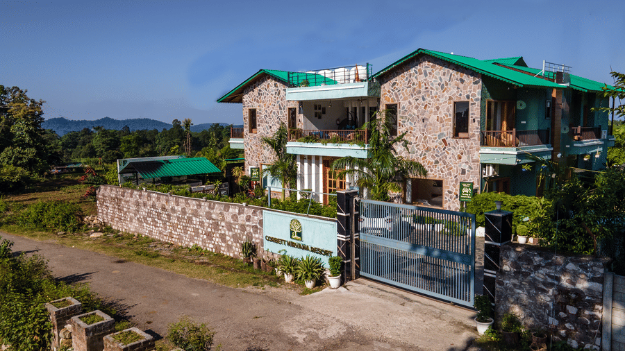 Day-time exterior view of the multi-storey, stone-clad main building of Corbett Nirvana Resort with green roof accents, seen from the gate.