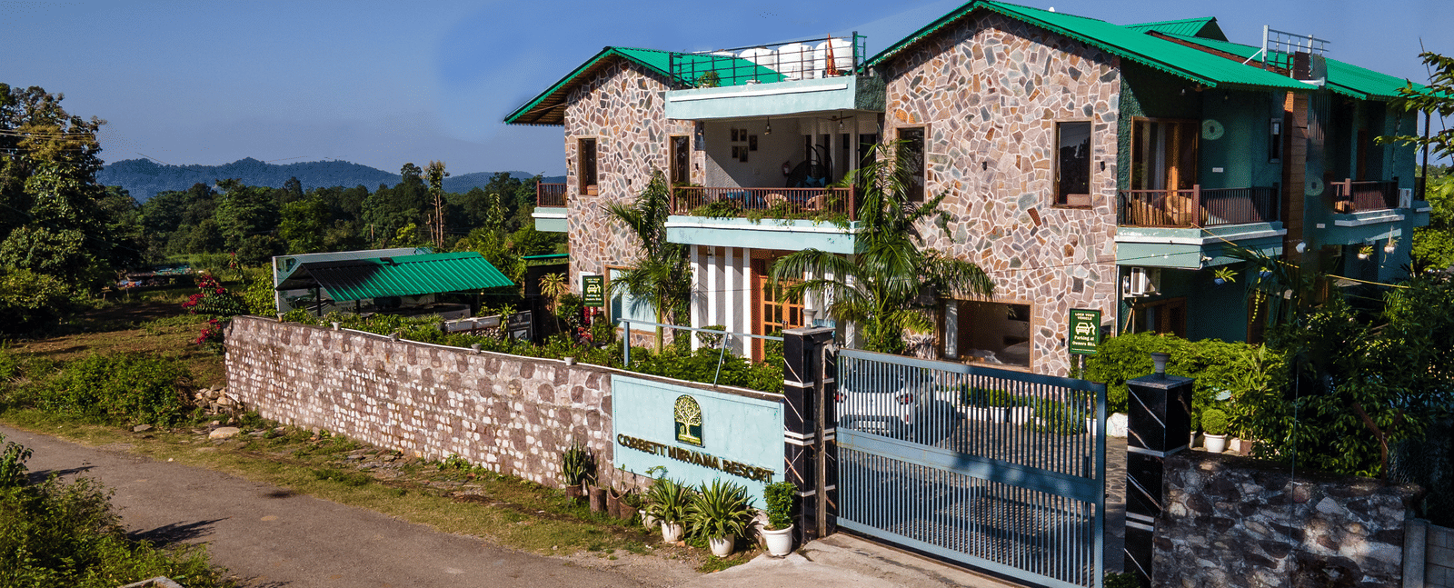 Day-time exterior view of the multi-storey, stone-clad main building of Corbett Nirvana Resort with green roof accents, seen from the gate.