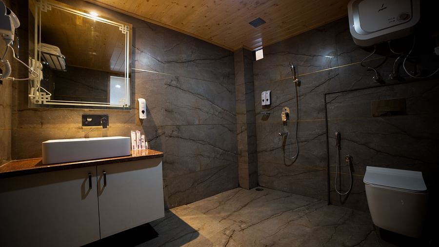  Modern bathroom with dark marble-patterned walls and flooring, featuring a wall-mounted sink with mirror, toilet, overhead water heater, and a walk-in shower area with mounted fixtures and a handheld bidet - Yashshree Lachen.