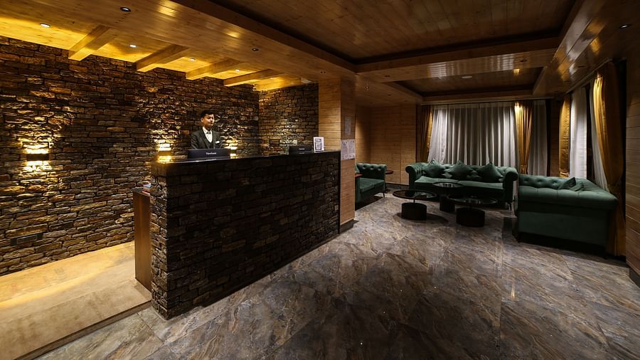 Modern hotel reception area with a dark stone front desk, warmly lit wooden ceiling, marble flooring, and green velvet seating in a cosy waiting lounge - Yashshree Lachen.