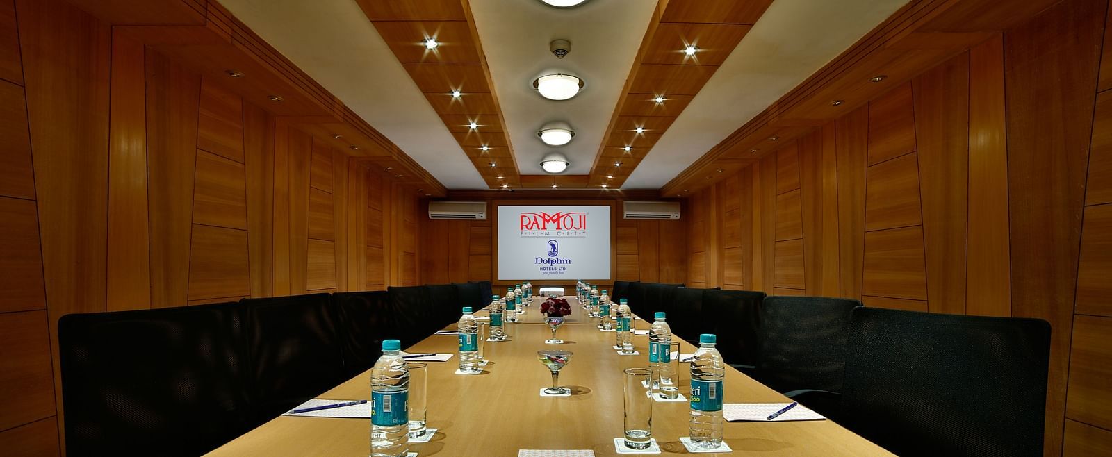 A sleek boardroom with a wooden table, leather chairs, and a screen for presentations. - Tara, Comfort Hotel in Hyderabad - Ramoji Film City.