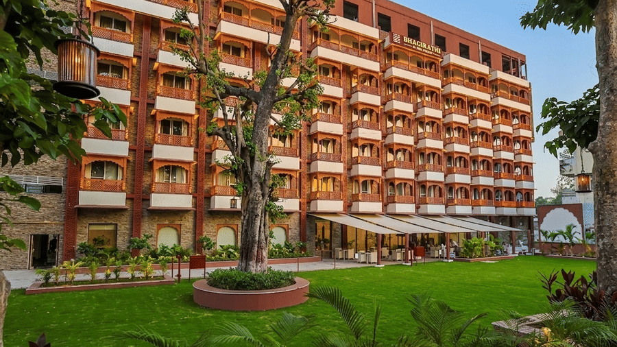 Exterior view of a multi-story building featuring wooden balconies, a manicured green lawn, and a shaded outdoor seating area - Hotel Bhagirathi