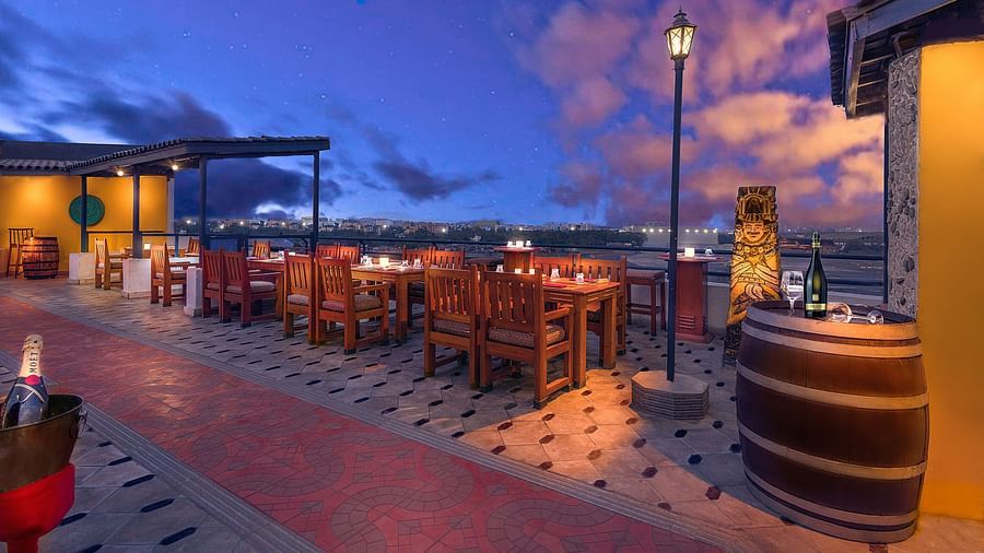 Rooftop dining area at dusk with wooden tables lit by candles, offering a scenic view of the city skyline under a vibrant, cloud-streaked sky.