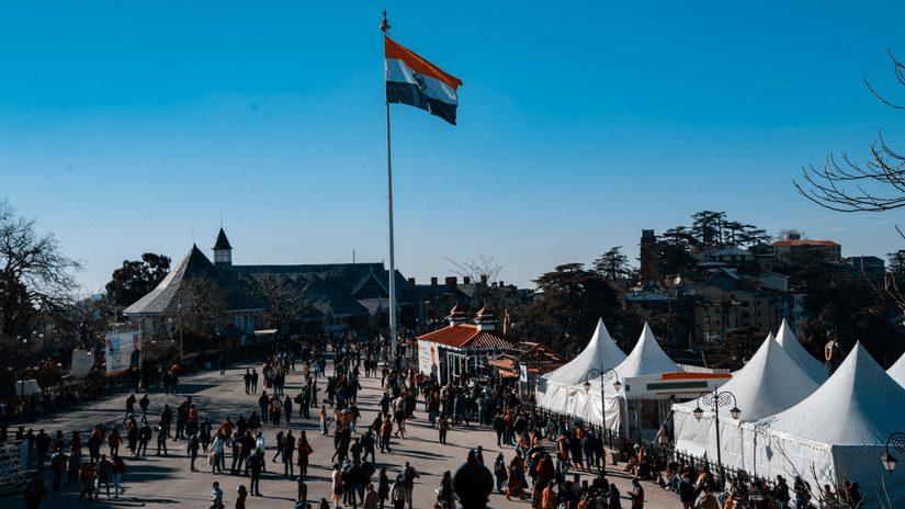 An overview of the Ridge in the morning with an Indian flag on a pole, many people walking on the street and shops lined on the sides.