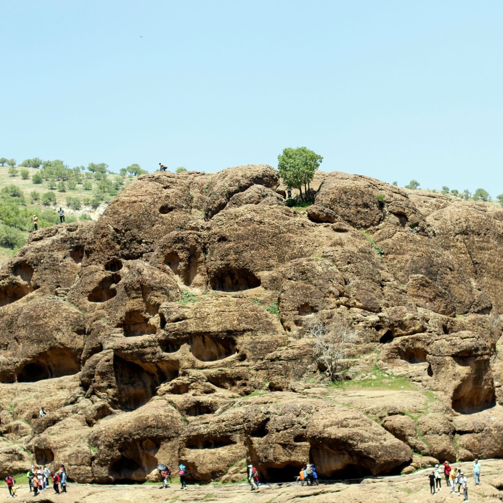 A boulder hill around a desert area with people walking below on a sunny day
