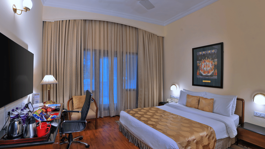 A room with a large bed, spreads, pillows, a TV, a chair, a table, and windows. | Clarks Varanasi
