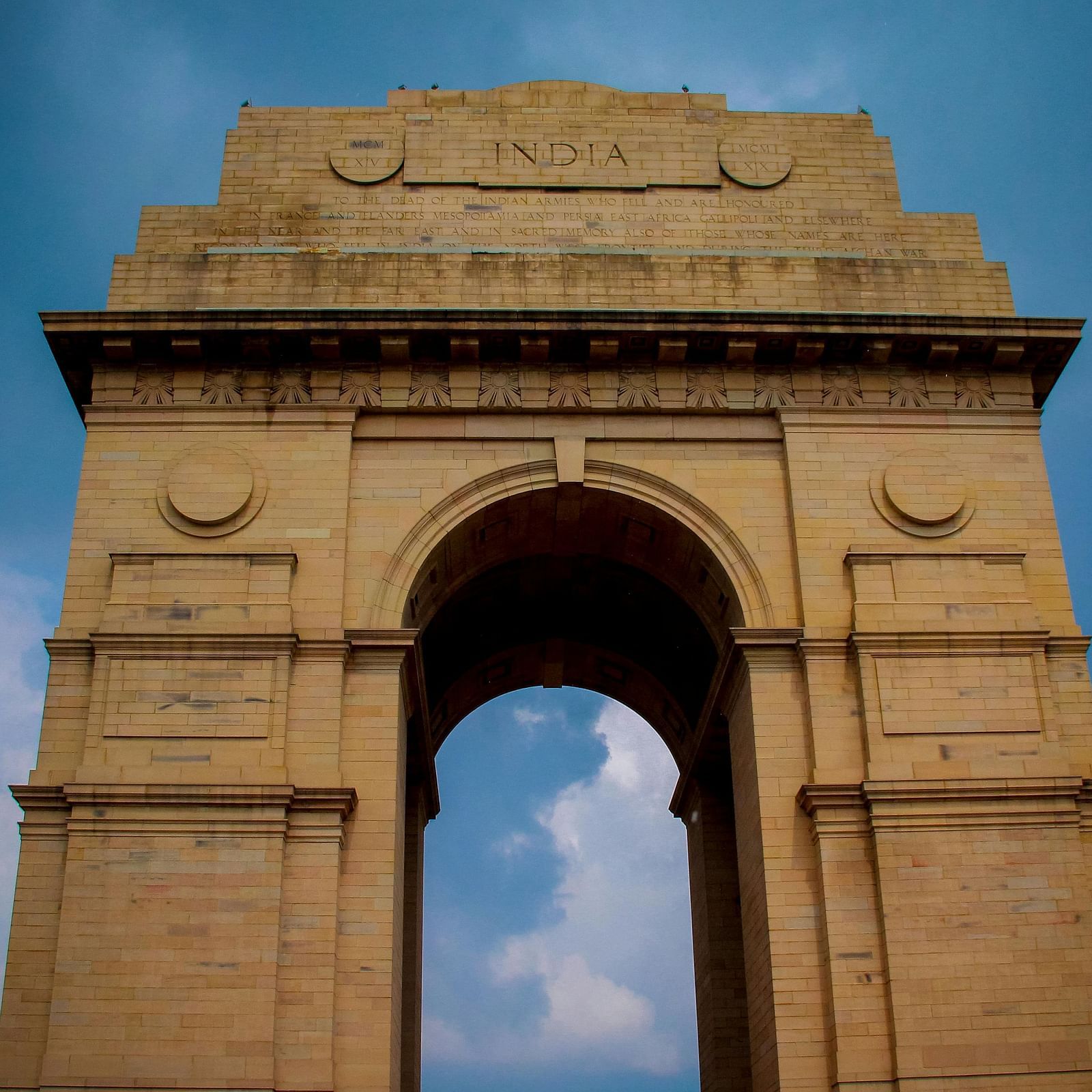 The India Gate, a large, arched war memorial made of light brown stone, stands against a dramatic blue sky with scattered clouds.