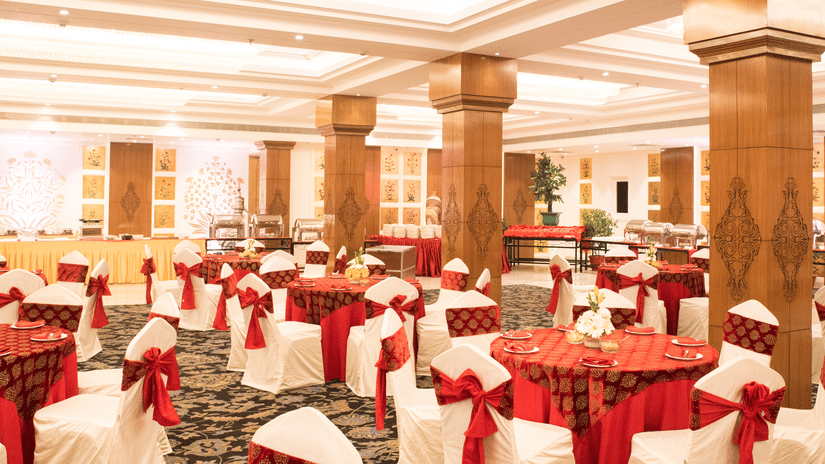 A grand banquet hall with high ceilings, large pillars, and elegant red and gold decor, with a lavish dining setup for a wedding or event - Clarks Avadh, Lucknow