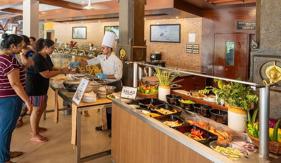 Guests being served at a live chaat counter by a chef in uniform, with a colourful salad bar in the foreground and others queuing in a warmly lit buffet setting at Uday Samudra Leisure Beach Hotel.