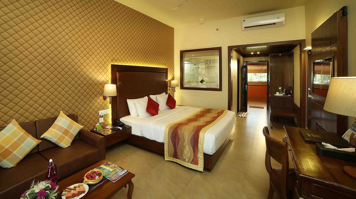 The Ocean View Room at Uday Samudra Leisure Beach Hotel & Spa, Kovalam,  features a king-size bed with decorative pillows and a patterned bed runner, a brown sofa with cushions, a wooden desk and chair and food placed on the coffee table behind.