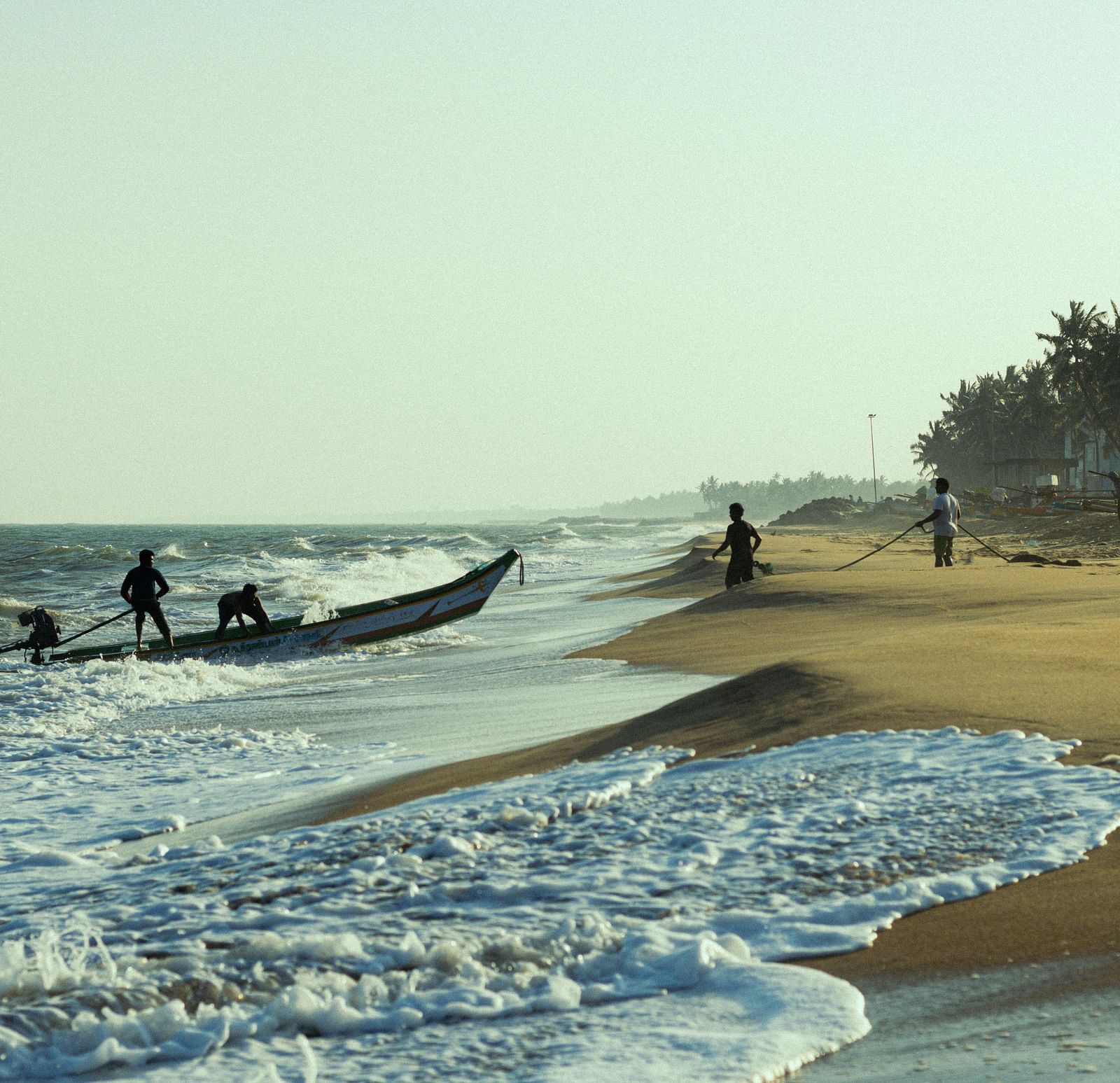 A group of people pulling a fishing boat into the ocean at the beach in Pondicherry, with palm trees visible along the shoreline in the background.