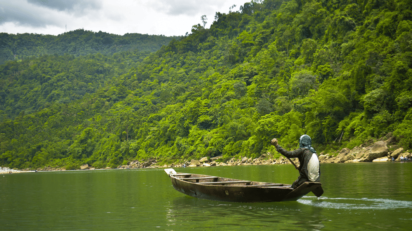 Lone boatman in a wooden canoe rowing across a green river with dense forested hills rising behind under a cloudy Meghalaya sky.
